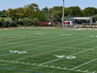 Show thumbnail preview A green, artificial turf football field marked with yard lines and numbers. The field is empty, with goalposts visible in the distance. Trees and a few vehicles, including a red truck, can be seen in the background under a clear blue sky.