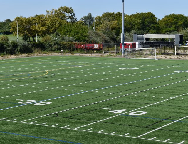 Show thumbnail preview A green, artificial turf football field marked with yard lines and numbers. The field is empty, with goalposts visible in the distance. Trees and a few vehicles, including a red truck, can be seen in the background under a clear blue sky.