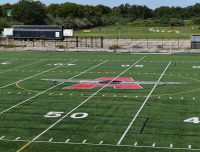 Show thumbnail preview A football field with a large red and black logo at the center, marked with white yard lines and hash marks. The number 50 is visible in the foreground. Trees and a few trailers are in the background under a clear sky.