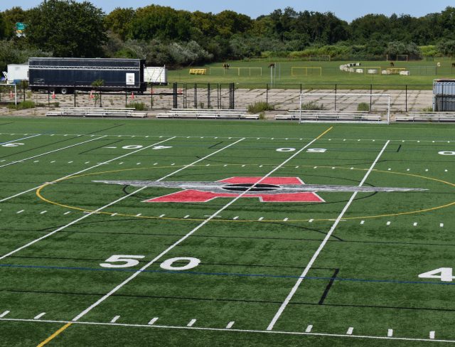 Show thumbnail preview A football field with a large red and black logo at the center, marked with white yard lines and hash marks. The number 50 is visible in the foreground. Trees and a few trailers are in the background under a clear sky.